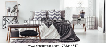 Wooden bench in grey bedroom interior with patterned pillows on bed and plant on white nightstand