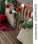 A wooden bench on a porch adorned with red and white pom-pom garland, patterned pillows, and a warm plaid blanket, creating a festive and cozy holiday atmosphere.