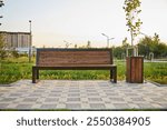 wooden bench paired with a metal trash bin in tranquil park. Warm light as the sun sets, casting long shadows across the checkered pavement. Grass and young trees, peaceful ambiance. city improvement