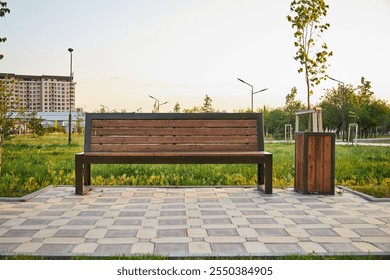 wooden bench paired with a metal trash bin in tranquil park. Warm light as the sun sets, casting long shadows across the checkered pavement. Grass and young trees, peaceful ambiance. city improvement - Powered by Shutterstock