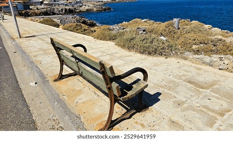 A wooden bench with rusty armrests and legs sits on a sandy-colored pavement, offering a view of the turquoise sea, creating a charming coastal scene. - Powered by Shutterstock