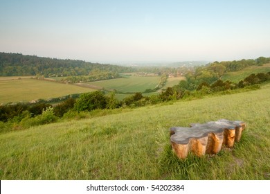 A wooden bench shaped like a leaf is in the foreground of a landscape shot of the North Downs in Surrey, UK.
