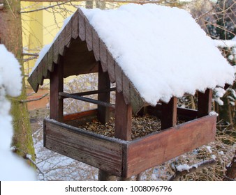 A Wooden Bird Feeder Stands On A Branch Nearby Yellow House. A Wooden Feeder Roof Is Full Od Snow And Some Bird Seed Is Inside. It Is Concept Of Winter And Bird Care (feeding) In The City Or Village.