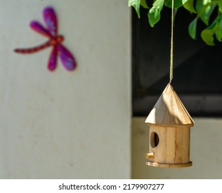 Wooden Birdhouse Hanging From A Tree Branch In The Garden With The Colorful Silhouette Of A Decorative Dragonfly On The Wall