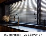 Wooden blinds black color closeup on the window. Bamboo slats 50mm wide. Venetian wood blinds in the kitchen. Black tapes. Sink with copper faucet near the window. Round vase is on the windowsill.