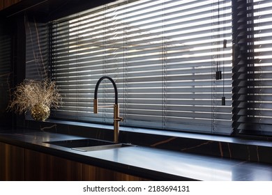 Wooden blinds black color closeup on the window. Bamboo slats 50mm wide. Venetian wood blinds in the kitchen. Black tapes. Sink with copper faucet near the window. Round vase is on the windowsill.