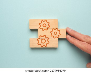 Wooden Blocks With A Gear Icon On A Blue Background, One Hand Puts The Wooden Blocks Together