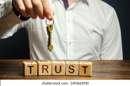 Wooden Blocks With The Word Trust And Keys In The Hands Of A Businessman. Entrust Apartment, Car, Property And Real Estate. Credit Rating. Guarantee For Rent. Reliable Partner