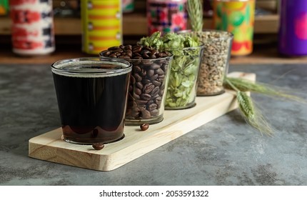 Wooden Board For Flight Set Craft Beer Tasting. Coffee Stout Or Porter With Ingredients. Bar With Different Cans In The Blurred Background. 