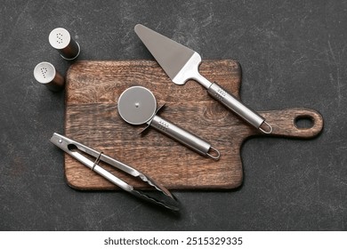 Wooden board with stainless steel pizza cutter, spatula, tongs and spices on dark background - Powered by Shutterstock