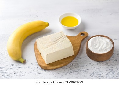 Wooden Board With Tofu, Banana, Rice Flour And Vegetable Oil. Ingredients For Vegan Cheesecakes Or Other Vegan Pastries. On A Light Blue Table