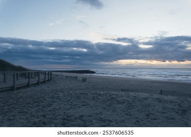 A wooden boardwalk leads to a beach with cloudy skies and ocean waves. The beach is sandy and the sky is a soft blue. There are clouds in the sky. The sun is setting and the sky is a soft pink. - Powered by Shutterstock