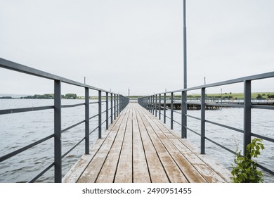 A wooden boardwalk with a metal railing crosses calm water under a clear sky, creating a peaceful area for a relaxing outdoor walk by the tranquil lake surrounded by tranquil nature - Powered by Shutterstock