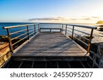 A wooden boardwalk overlooking the ocean. The boardwalk is empty and the sky is blue. The sun is setting and the water is calm