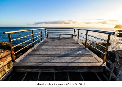 A wooden boardwalk overlooking the ocean. The boardwalk is empty and the sky is blue. The sun is setting and the water is calm - Powered by Shutterstock