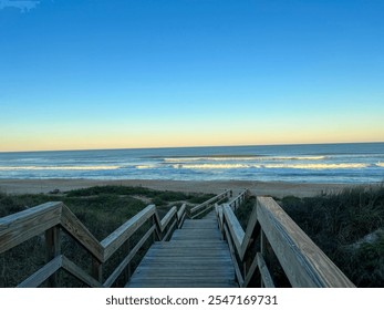 A wooden boardwalk stretches towards a peaceful sandy beach, leading to the shimmering ocean waves under a vast, clear blue sky with golden light on the horizon. - Powered by Shutterstock