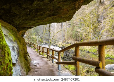 A wooden boardwalk winds through a scenic gorge in Bohemian Switzerland, Czechia. A large rock overhangs the path, offering shade as hikers explore the natural beauty of the area. - Powered by Shutterstock