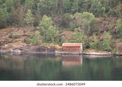 A wooden boat hut on the shore of a Norwegian fjord with reflections in the water - Powered by Shutterstock