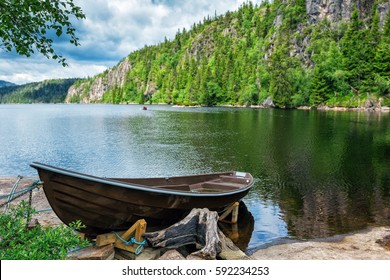 Wooden Boat On The Lake Bank On Forest Background And Blue Sky Reflecting In The Surface Of The River, Norway