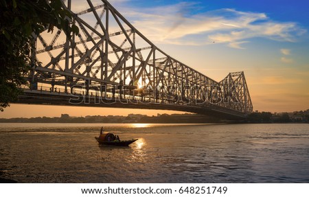 Wooden boat passing the historic Howrah bridge in silhouette at sunrise. Howrah bridge is a cantilever bridge on the river Hooghly and one of the busiest bridge in the world.