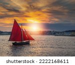Wooden boat with red color sail going into harbor. Galway city, Ireland. Popular local type boat called Galway hooker. Sport and hobby. City building silhouette in the background. Dramatic sunset sky.