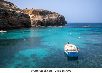A wooden boat with vibrant, sun-faded paint drifts on the clear, azure waters of the Maltese sea, framed by the rocky coastline and historic buildings under a clear, blue sky. - Powered by Shutterstock
