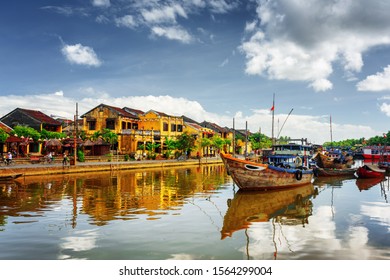 Holzboote auf dem Thu Bon River in Hoi An Ancient Town (Hoian), Vietnam. Geräumige, gelbe Häuser am Ufer, die sich im Fluss widerspiegeln. Hoi An ist ein beliebtes Reiseziel Asiens.
