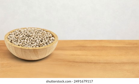 A wooden bowl filled with white peppercorns sits on a wooden surface against a white background. The peppercorns are natural, showcasing their smooth, round, pale beige appearance. - Powered by Shutterstock