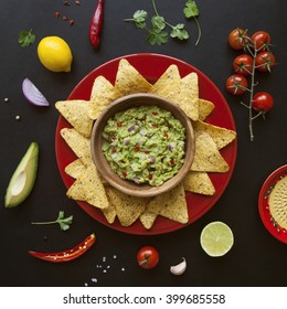 Wooden Bowl Of Fresh Homemade Guacamole, Its Ingredients And Nachos Chips On Black Background From Above