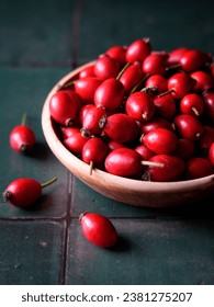 Wooden bowl full of red rosehips on a green tile background.