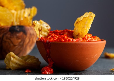 A Wooden Bowl Of Potato Chips With A Terra Cotta Bowl Of Salsa Or Hot Pepper Dipping Sauce. A Chips Has Been Dipped In. Some Mess And Spilling On Dark Moody Background. A Late Night Snack, Appetizer