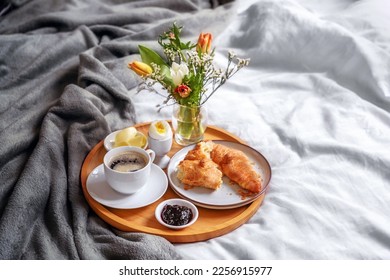 Wooden breakfast tray with croissant, coffee, jam, egg and a fresh flower bouquet served on bed with light linen and gray blanket, holiday or weekend morning, copy space, selected focus