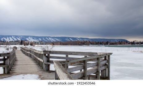 A wooden bridge in Lighthouse Point, Collingwood, crosses a portion of Georgian Bay while overlooking the Blue Mountain Ski hills from a distance, with the frozen water beside it