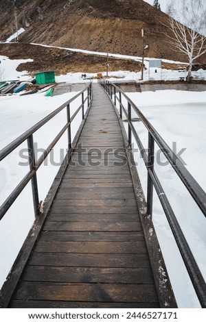A wooden bridge with a metal handrail crosses over a snowy field, contrasting with the white landscape. The composite material fence blends with the sky