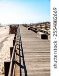 A wooden bridge stretches through a sandy dune landscape with railings, benches, and shadows. People walk on the bridge under a blue sky.