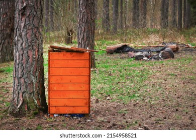 Wooden brigh orange trash bin in forest resting spot between tree trunks on green grass in spring