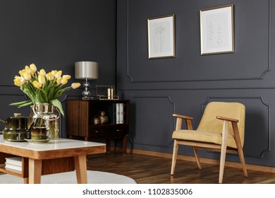 Wooden cabinet in the corner of a dark living room interior with wainscoting on the wall with posters next to a yellow armchair and table with tulips. Real photo