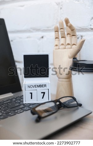 Wooden calendar with date November 17 and laptop, coffee cup, eyeglasses, wooden hand on table against brick wall background. Deadline, planning, business concept
