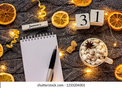 Wooden calendar January 31st Cup of cocoa with marshmallow, empty open notepad with pen, dried oranges, light garland on grey knitted background. Top view Flat lay Mockup