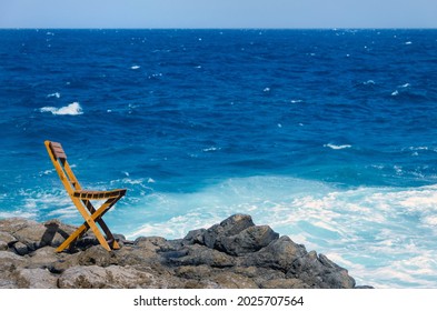 
Wooden Chair On The Edge Of The Sea In A Rocky Area While The Waves Are Beating And The Sky Is Blue With Some Clouds