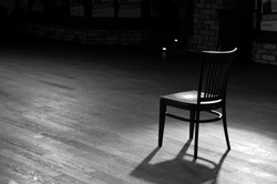 A Wooden Chair Standing In The Middle Of An Empty Stage Under The Light Of Soffits, In Black And White