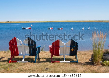 Wooden chairs along the shore and boat decorations on the water painted in the colors of the French Acadian flag on the coast near Bouctouche New Brunswick in the Canadian Maritime Provinces