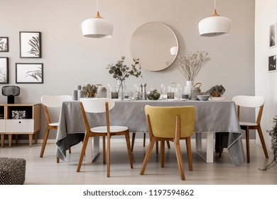 Wooden Chairs At Table With Tableware In Dining Room Interior With Lamps And Round Mirror. Real Photo