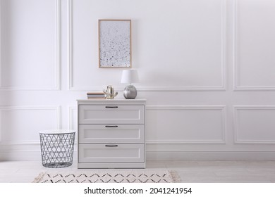 Wooden chest of drawers with books and lamp near white wall in room, space for text. Interior design