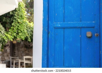 Wooden Closed Blue Old Door With Rusty Handle And Keyhole. Greek Island Vintage Entrance, Blur Traditional Cafe, Table Chairs, Plants Background.