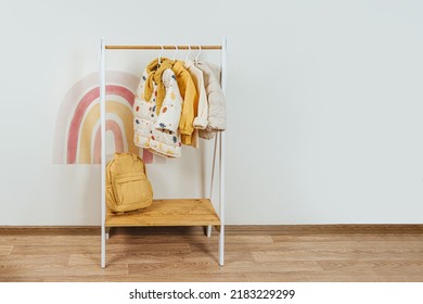 Wooden Clothing Rack With Children's Autumn Outfits. Dresses, Sweaters And Jacket In Kids Room. Montessori Wardrobe. Nursery Storage Ideas. 