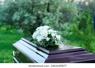 Wooden coffin covered with heavy lid with bunch of white flowers standing against green trees and bushes on cemetery before burial