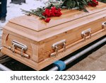 A wooden coffin with flowers on the lid stands above a dug grave in the cemetery. A closed coffin with red roses on it. Funeral ceremony at the cemetery in winter. Farewell ceremony and burial.