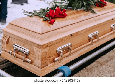 A wooden coffin with flowers on the lid stands above a dug grave in the cemetery. A closed coffin with red roses on it. Funeral ceremony at the cemetery in winter. Farewell ceremony and burial.