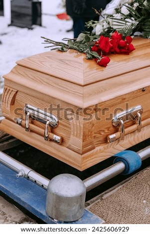 A wooden coffin with flowers on the lid stands above a dug grave in the cemetery. Red roses lie on the coffin. Funeral ceremony at the cemetery in winter. Farewell ceremony and burial. Close-up photo.